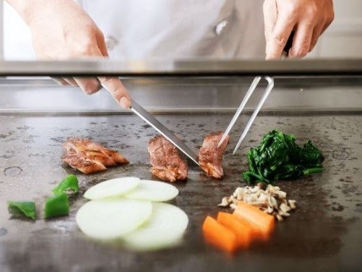 シェフが鉄板の上で肉を切り分けている。肉だけでなくピーマン、人参、玉ねぎなどの野菜も一緒に焼かれている。