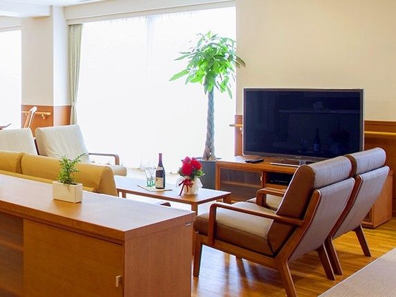 窓際の大画面テレビの向かいにテーブルがあり、テレビの正面にはソファが並んでいる。窓際には観葉植物、テーブルの上には小さな花が置かれている。