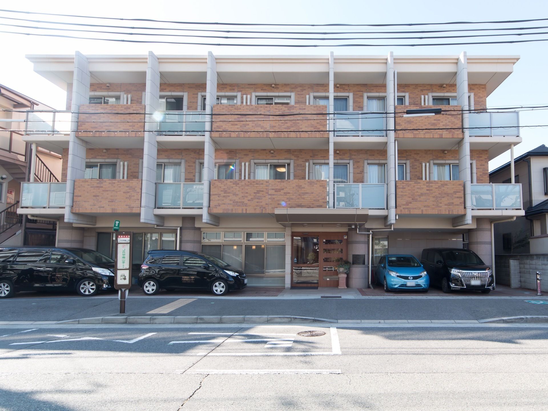 施設は3階建てで、茶色い色味の外壁で統一されている。正面には、黒色の車が3台と水色の車が1台停められている。
