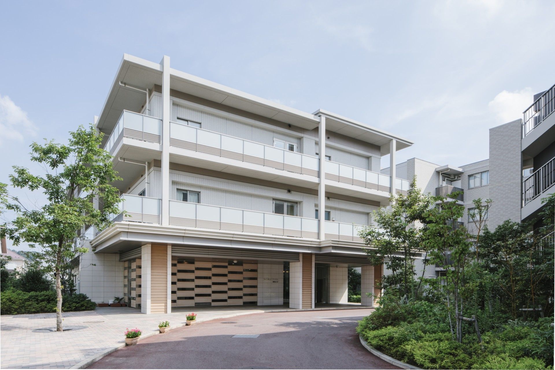 建物は４階建てで淡いベージュの外壁である。建物の周りには綺麗に手入れをされた木や花が植えられている。