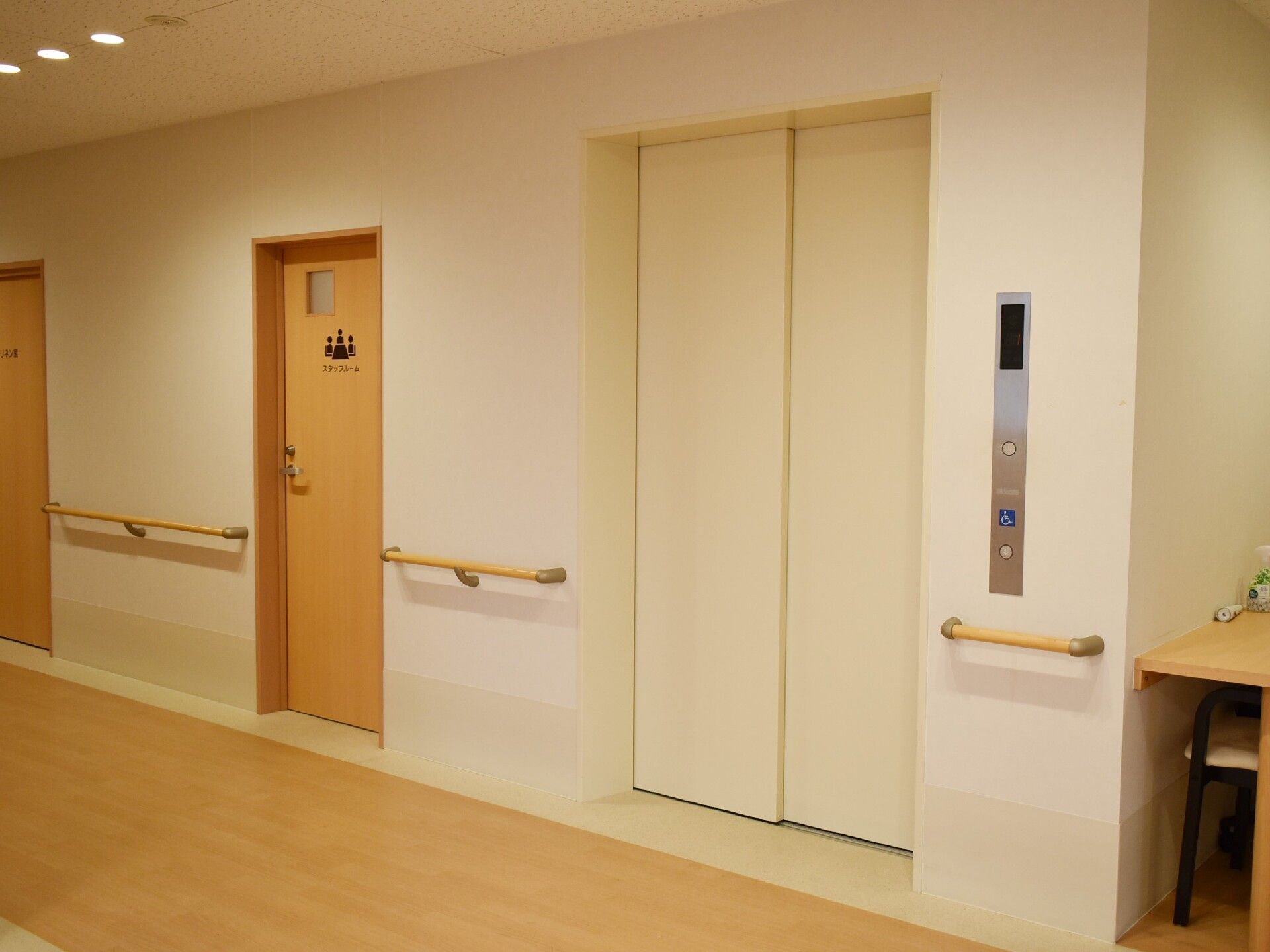 白い扉が付いたエレベーターが設置され、その左側には黒いロゴマークが描かれたドアがある。白い壁には手すりも付いている。