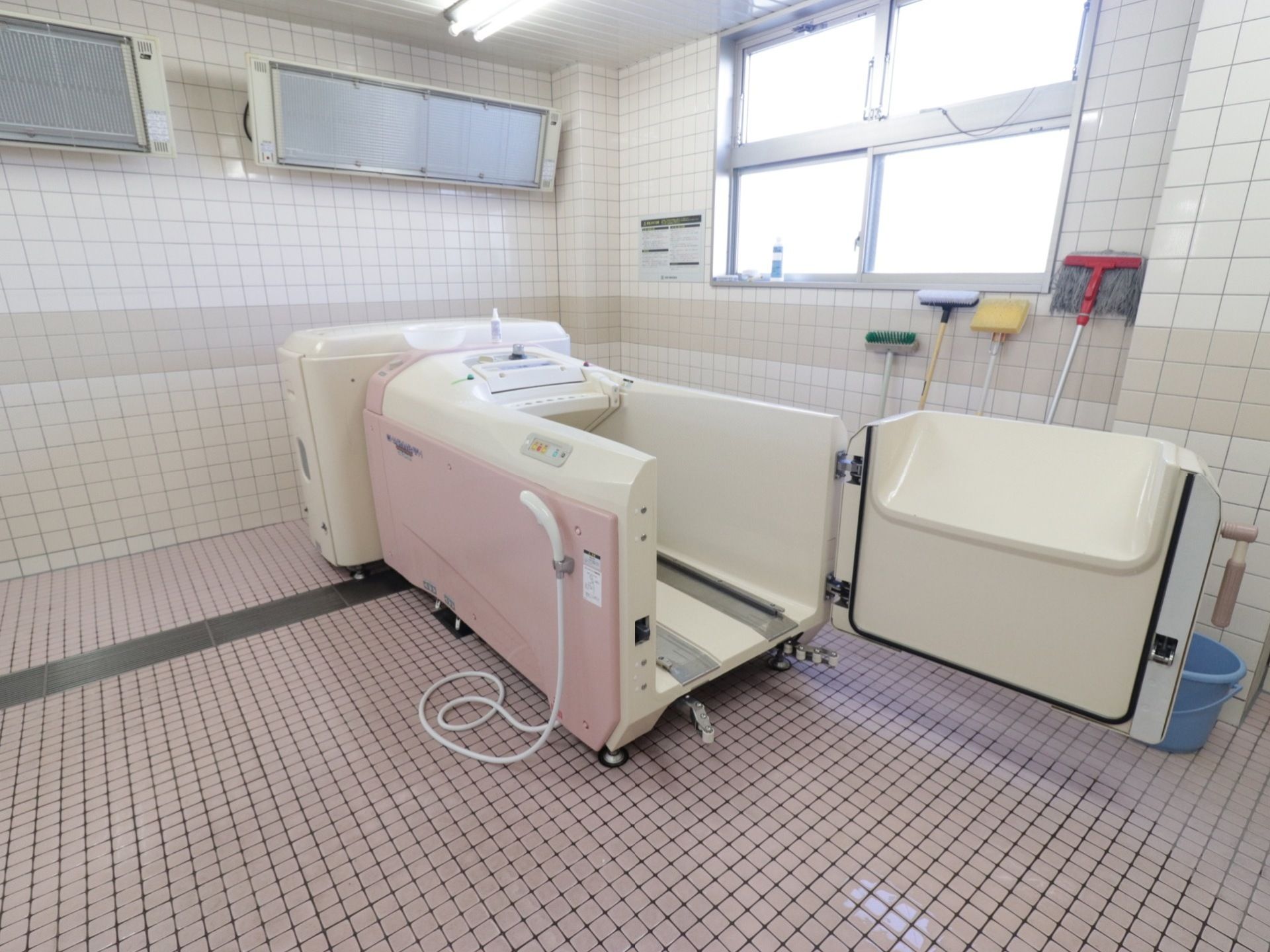 中央に介護入浴機器が置かれている。床はタイル敷きになっている。右側には窓があり、その手前には掃除道具が置かれている。