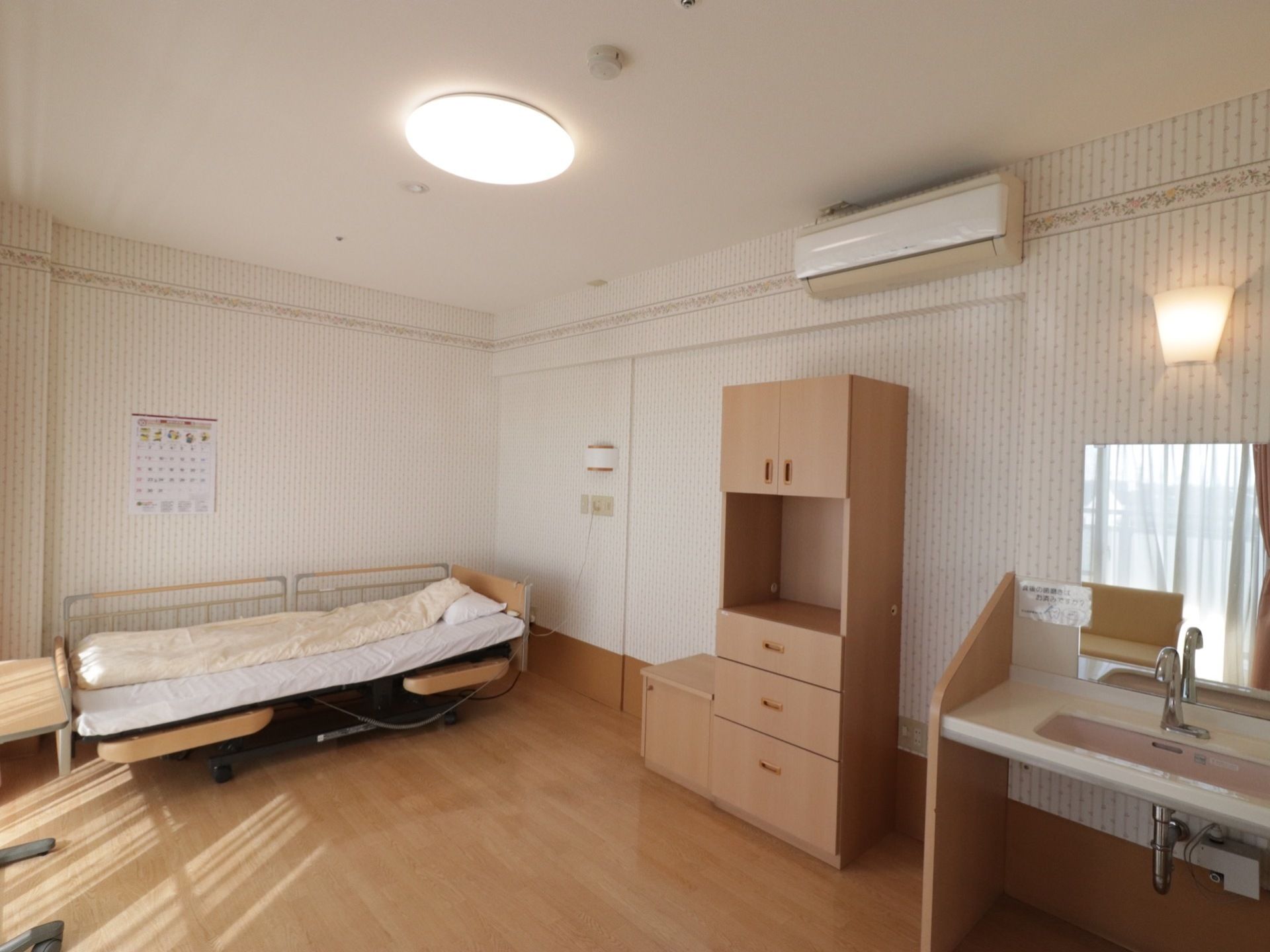 左側には介護用ベッドが置かれている。中央には収納棚があり、その上側にはエアコンがある。右側には洗面台がある。