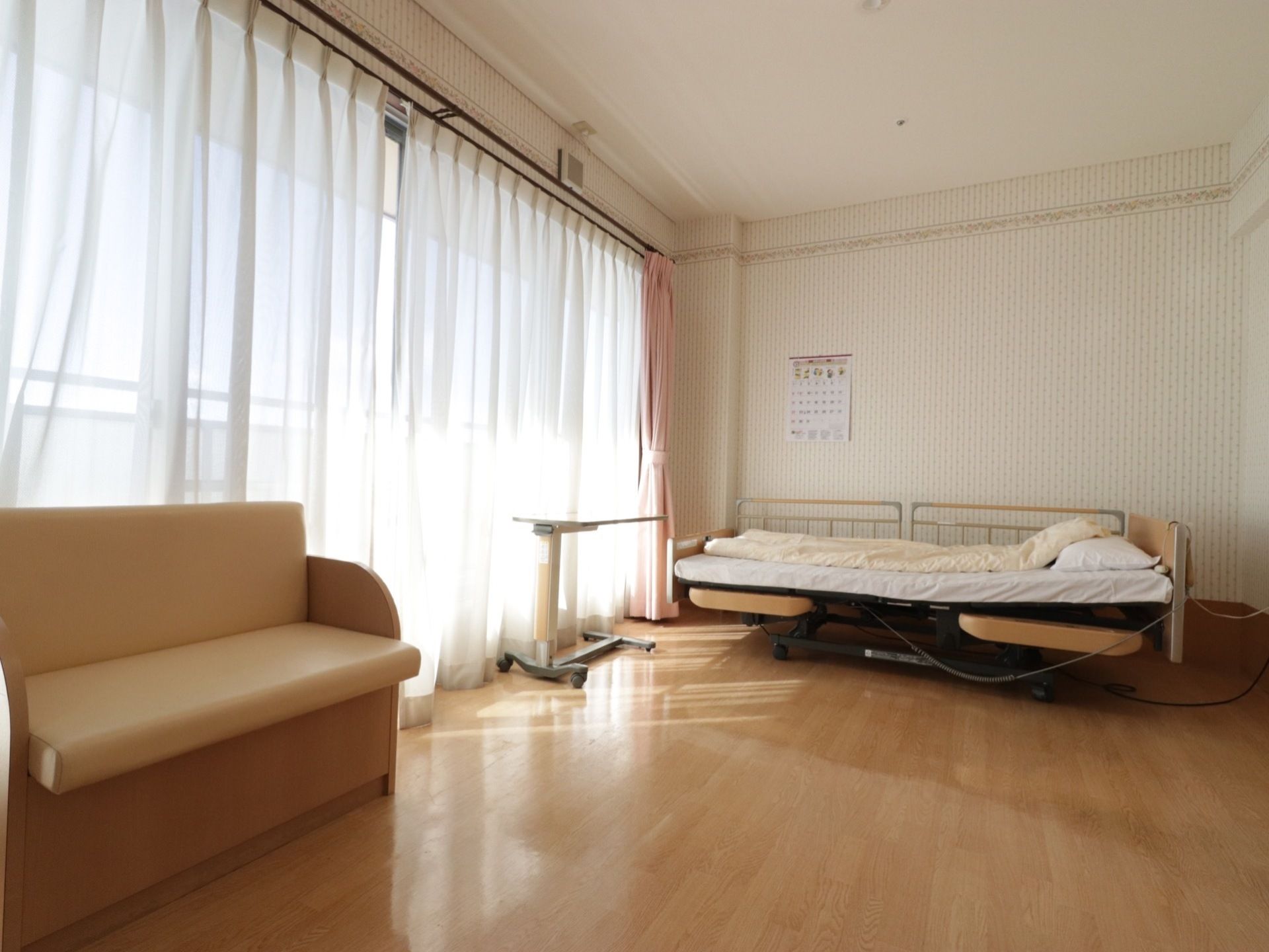 壁側に介護用のベッド、ベッドでも使用できる移動式の机がある。窓は大きく室内は明るい。ソファーも置いてある。