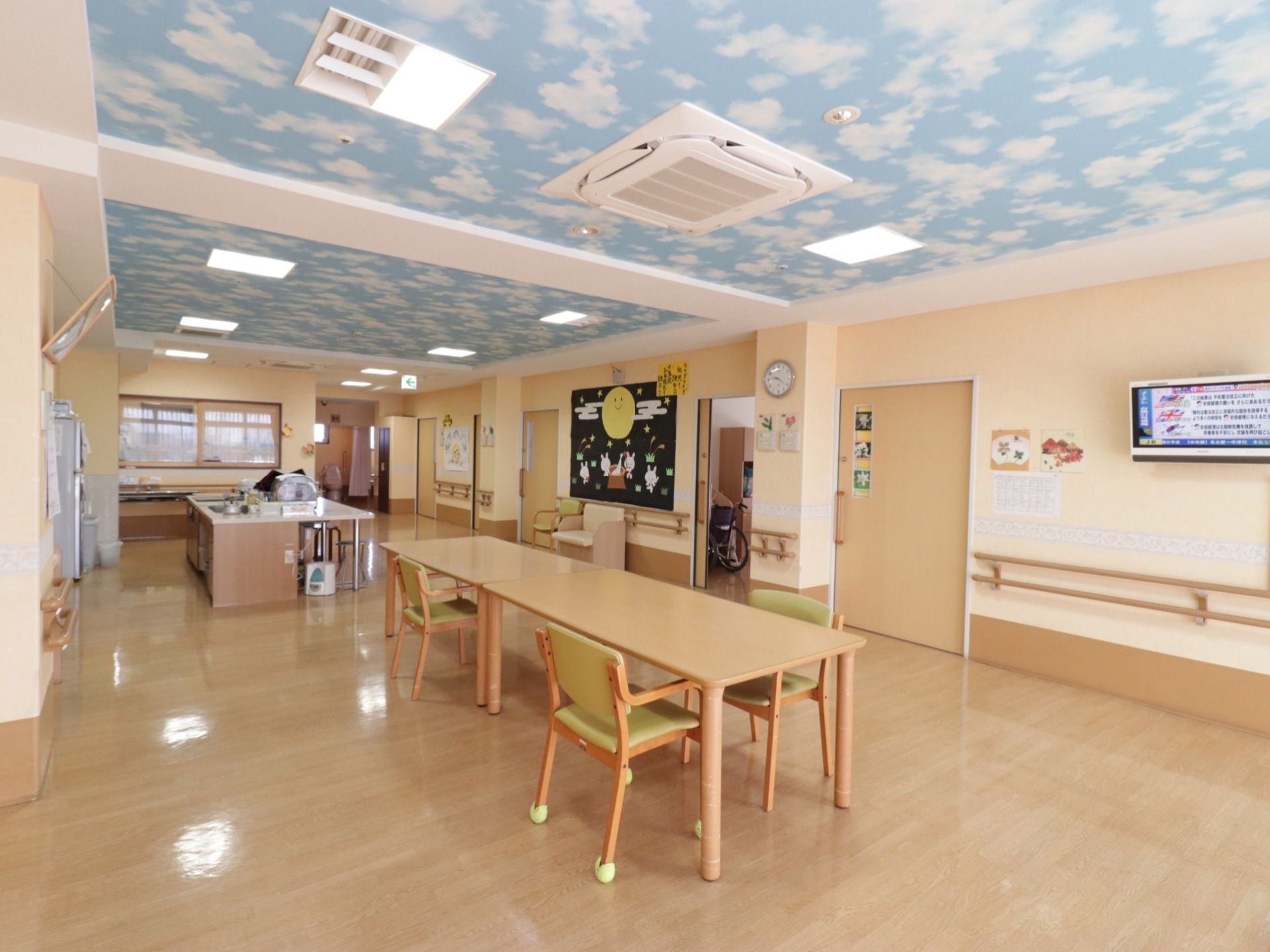 天井に空の壁紙が貼られている。中央には木製の机と椅子がある。壁には手すり、テレビの設置がある。キッチンも同じフロアにある。