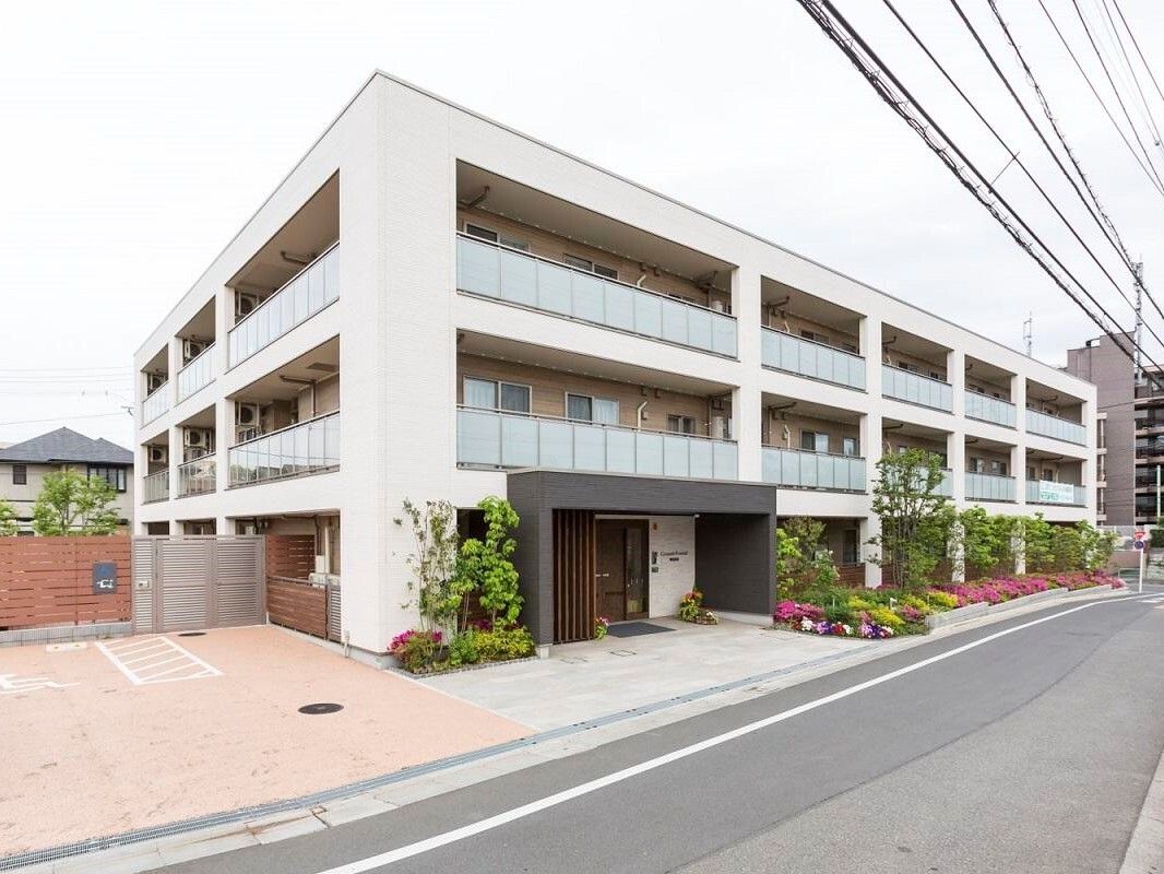 地上３階建ての施設は、ベージュ色の上品なデザインである。道路沿いには花壇があり、黄色い花や緑の草木が植わっている。