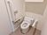 白い壁紙が清潔感のある、トイレの個室である。便座の上部には棚が設置されており、清掃用具などを収納できる。