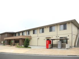 ベージュの外壁の２階建ての建物になっている。施設の前には駐輪場があり、その隣に自動販売機がある。玄関部分には屋根が設置されている。