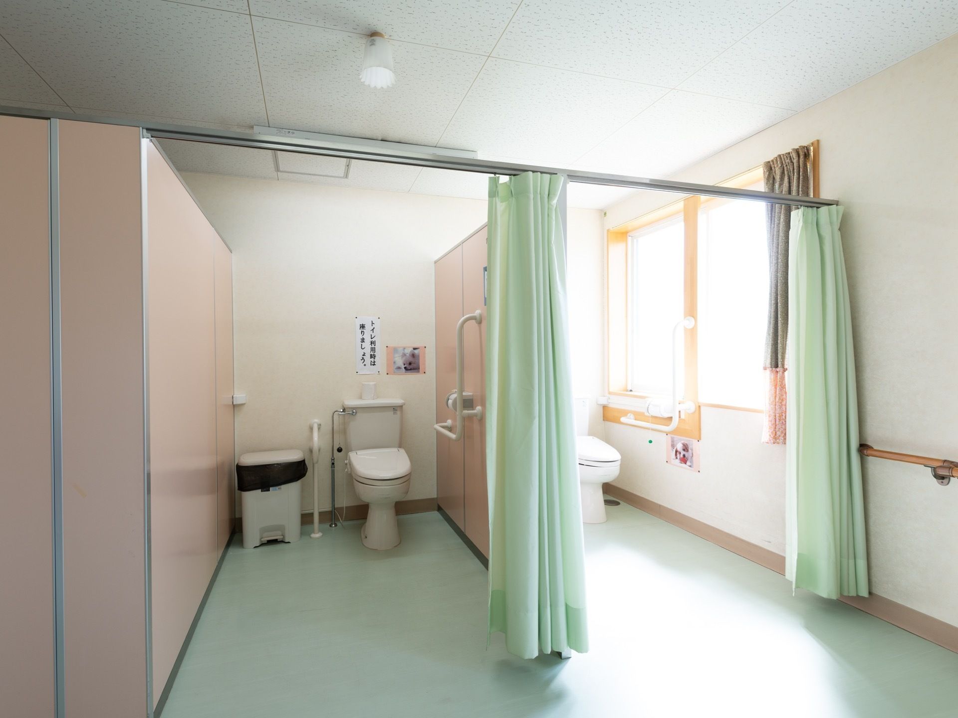 トイレには間仕切りのカーテンが設置されている。ゴミ箱が置かれており、壁には手すりやペーパーホルダーが取りつけられている。