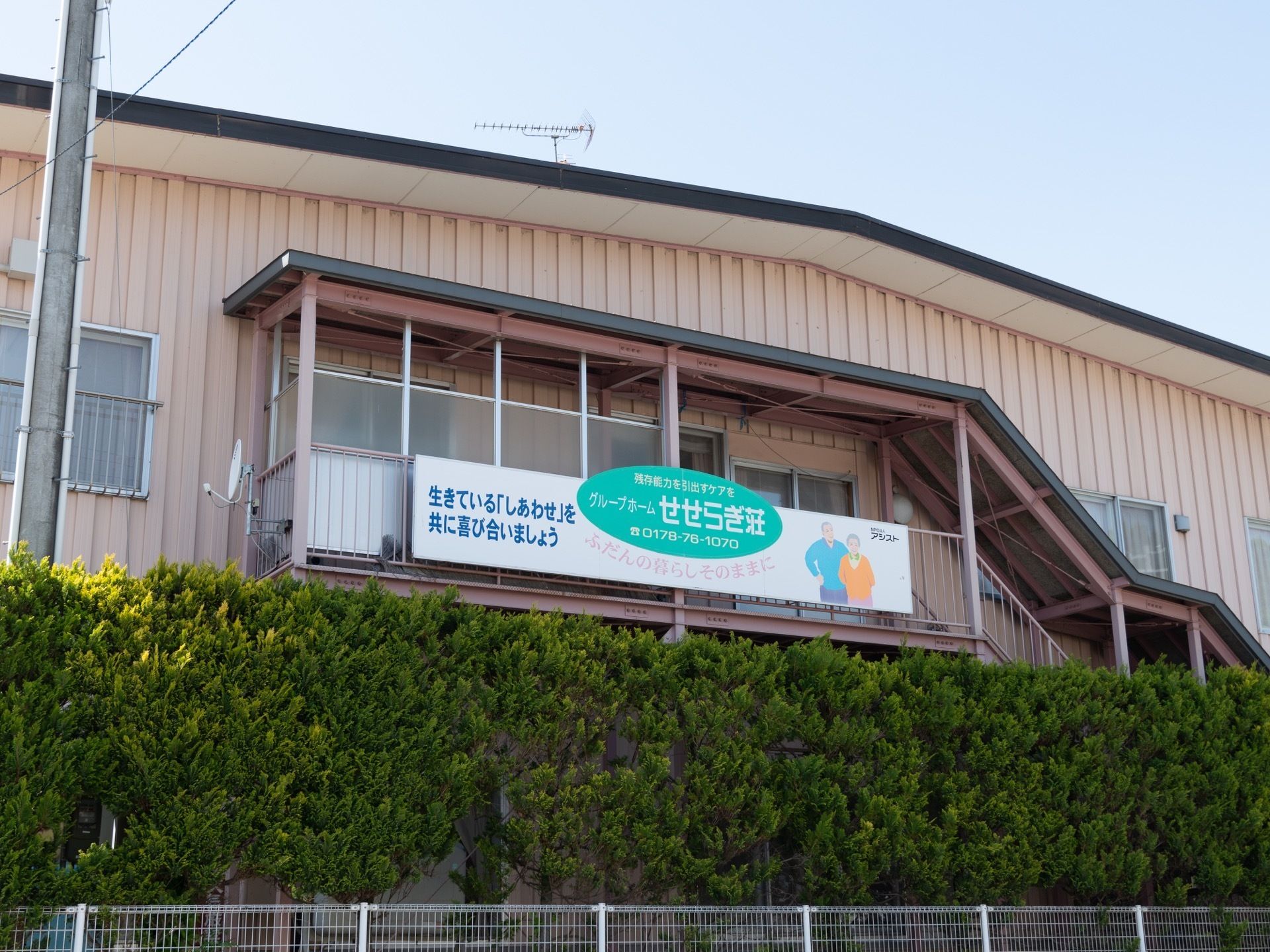 アットホームな雰囲気の施設外観である。施設名が記された看板が設置されており、道路から目立ちやすい。敷地内には植栽が施されている。