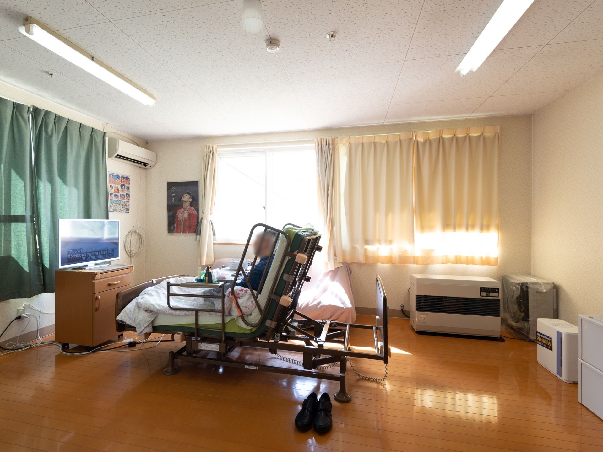 ２面の壁に大きな窓があって明るい居室である。入居者様が介護ベッドの半分を起こして、テレビを見ている。
