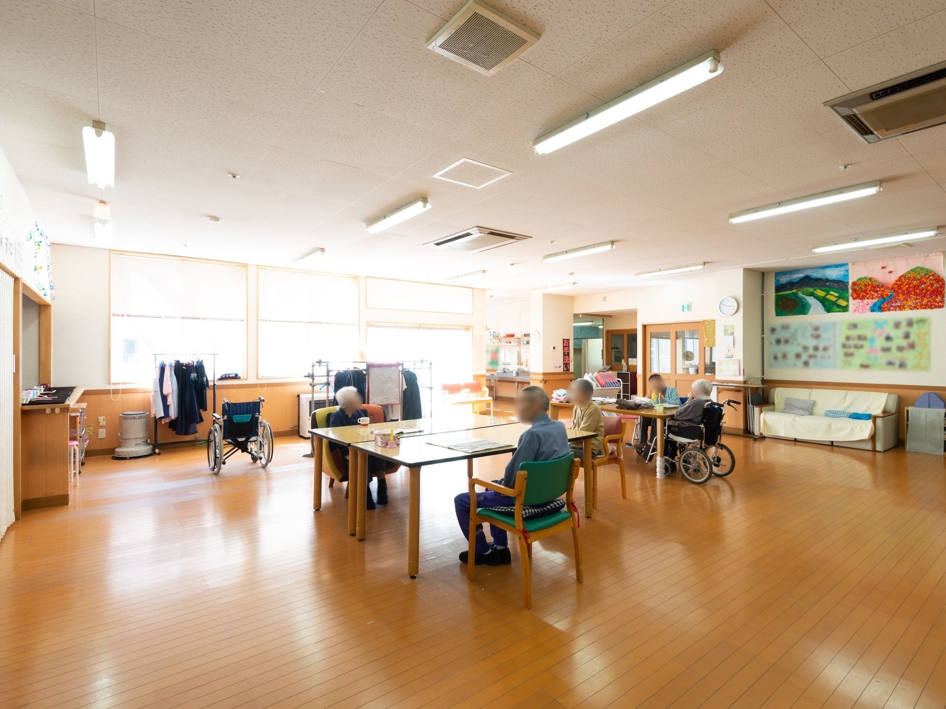 広い共有スペースに、テーブルと椅子が置かれている。入居者様が５名、椅子や車椅子に座ってテーブルについている。