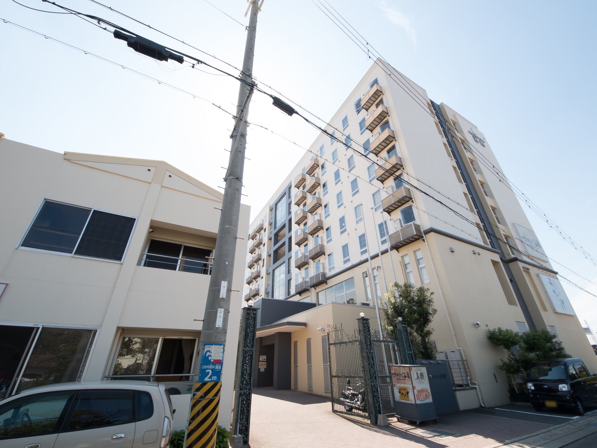 ８階建ての施設の隣に、２階建ての建物がある。目の前には電柱が立っている。軽四自動車が停められるスペースもある。