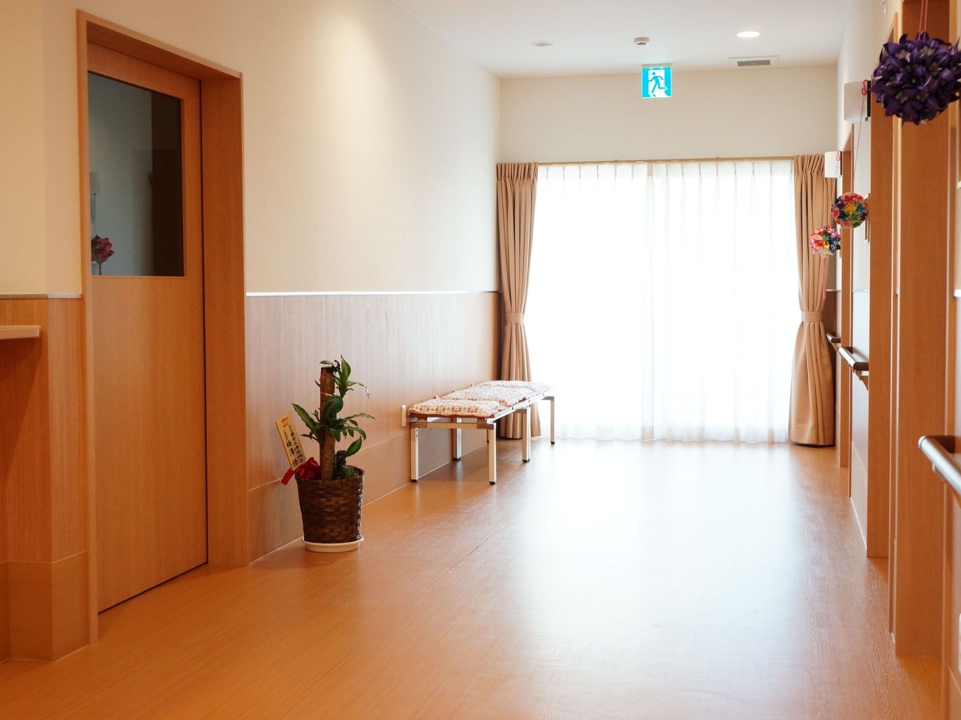 広々とした廊下。突き当たりの窓からは陽光が差し込んでいる。フローリングの床と木質系の壁は温かみがある。