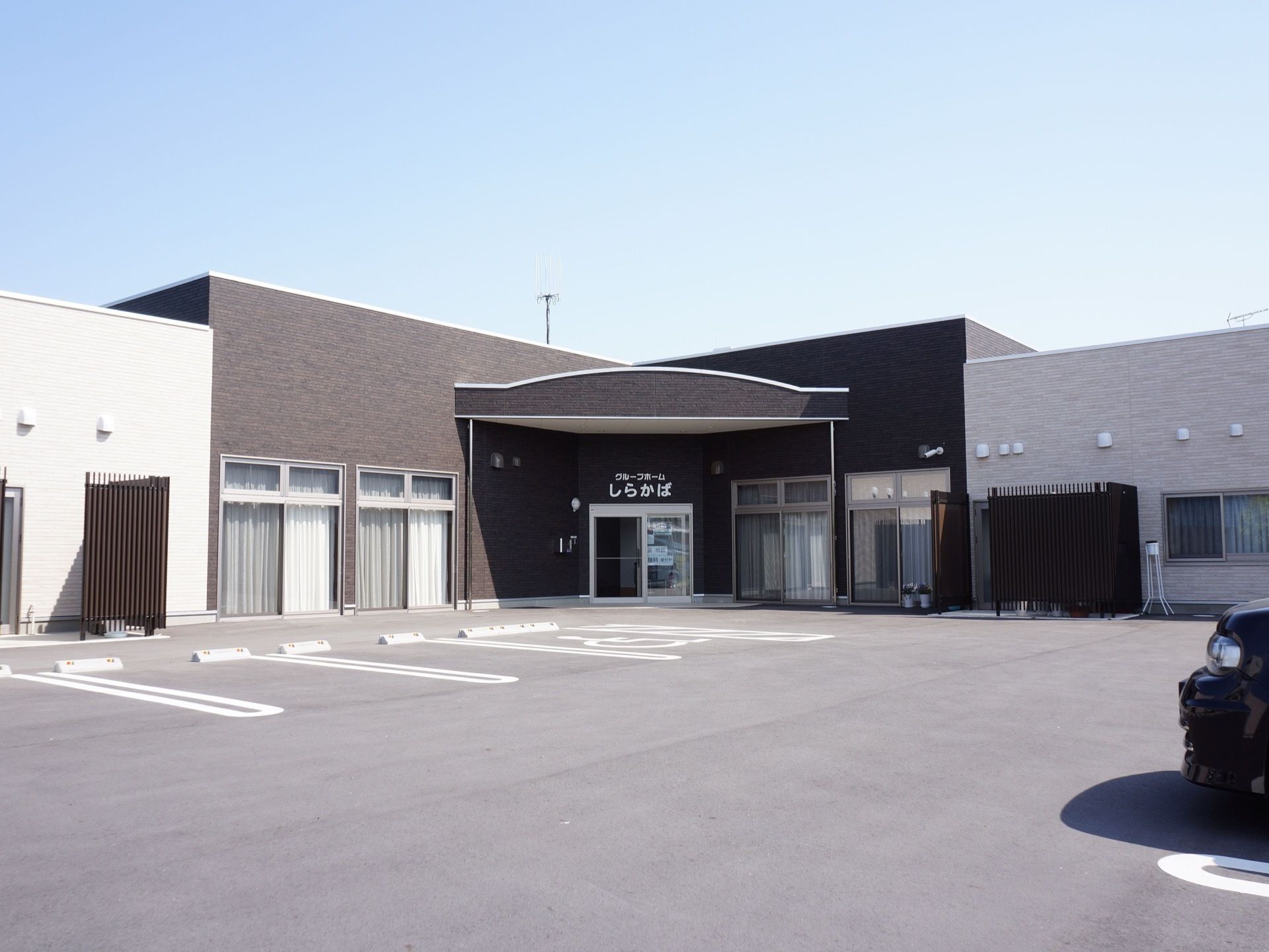 建物の周りには広々とした駐車場が広がっている。黒いレンガ調の出入口のある建物の両脇には、白い壁の建物が建っている。