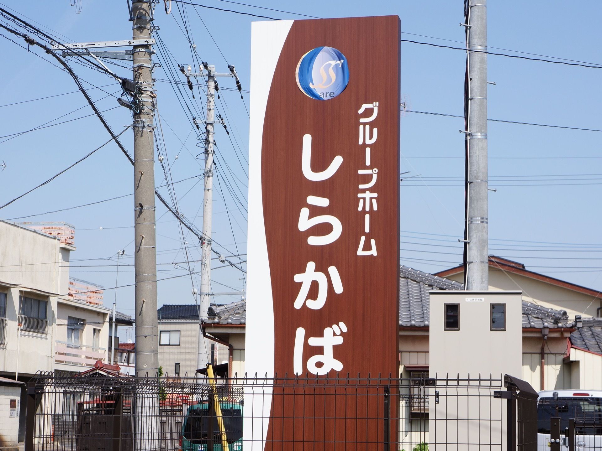 施設の名前が書かれた木目調の立て看板が住宅地の中に置かれている。立て看板の両脇には電信柱が立っている。