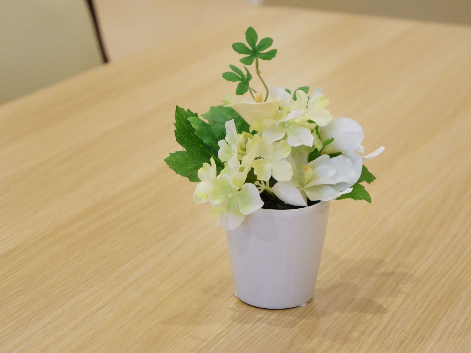 木目調のテーブルの上に黄色と白の造花の鉢が置かれている。緑色の葉が２枚、天井の方に向かって伸びている。