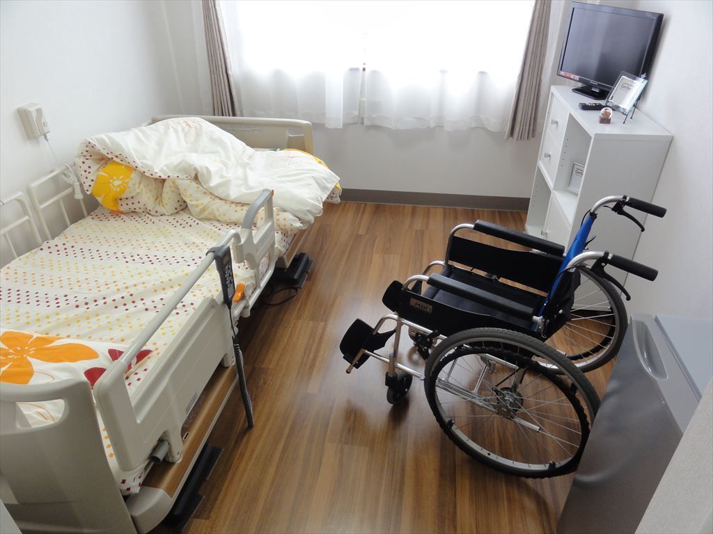濃い木目調の床の上にベッドやテレビ、車椅子がある居室