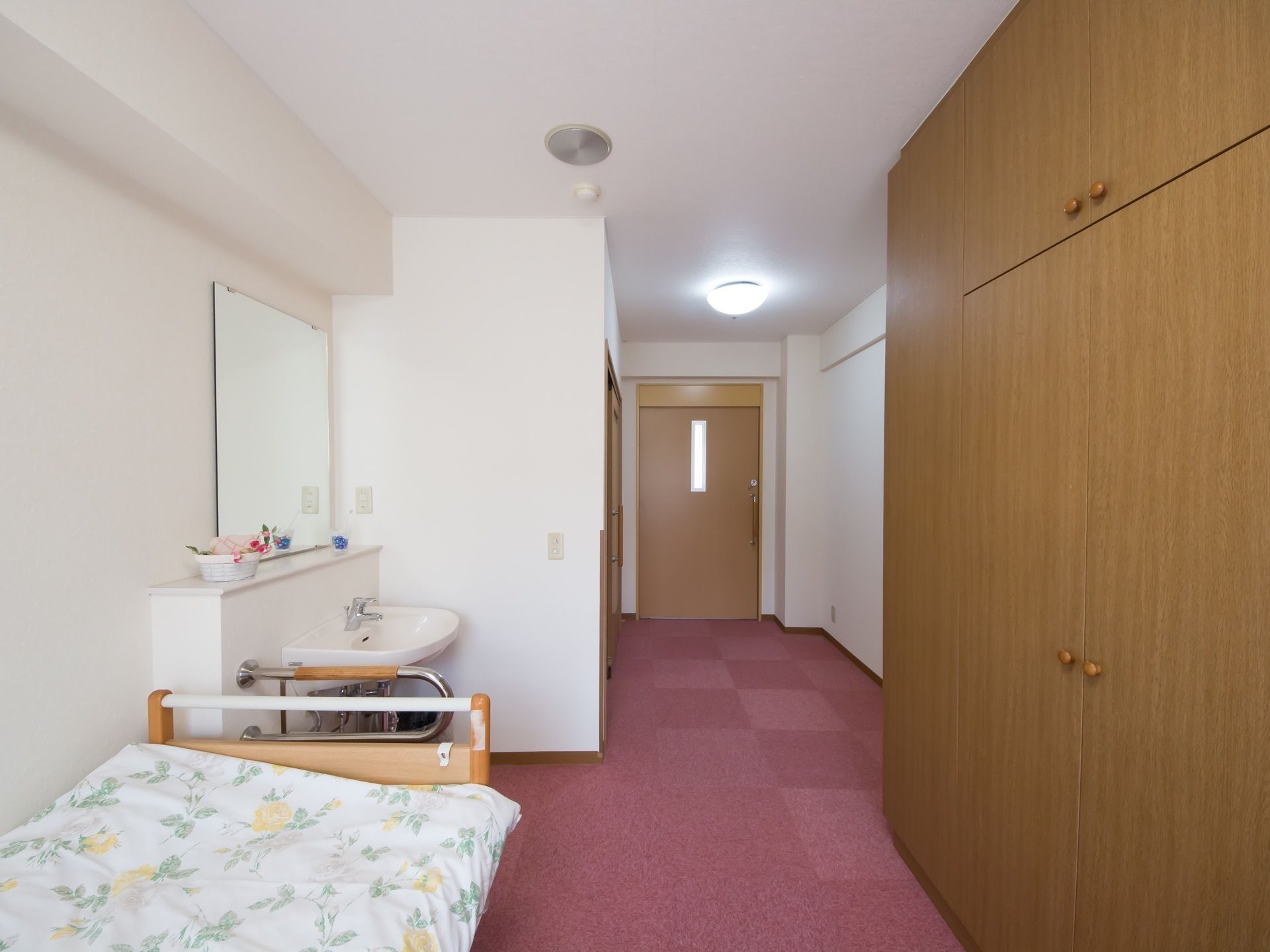 ピンクの床と白い壁の部屋に茶色の家具が置かれている。ベッドの横には大きなクローゼットが設けられている。