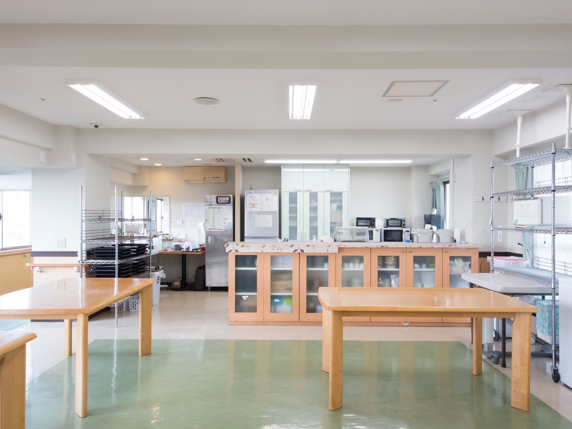 奥に食器棚や配膳台、電子レンジや冷蔵庫が置かれ、手前には机が並べられている。壁は白く、床はクリーム色と緑色になっている。