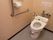 黄土色の壁に、グレイ色の床のトイレである。壁際には、茶色のエル型の手すりを設置し、便器の後部には、注意書きの紙を貼っている。