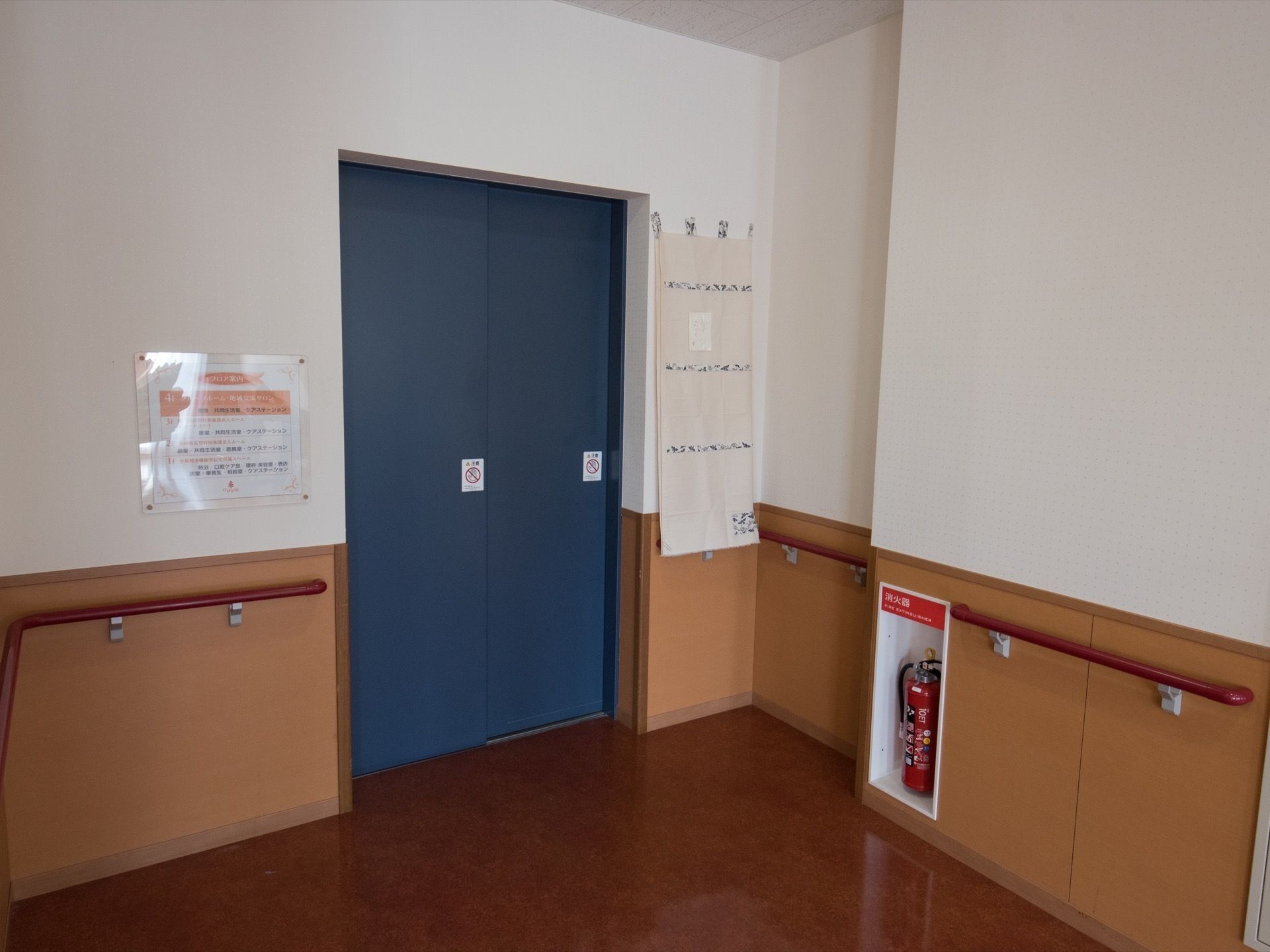 白色と茶色の壁に焦げ茶色の床の内装である。紺色のドアのエレベーターを備え、壁際に、消火器を設け、手すりを設置している。
