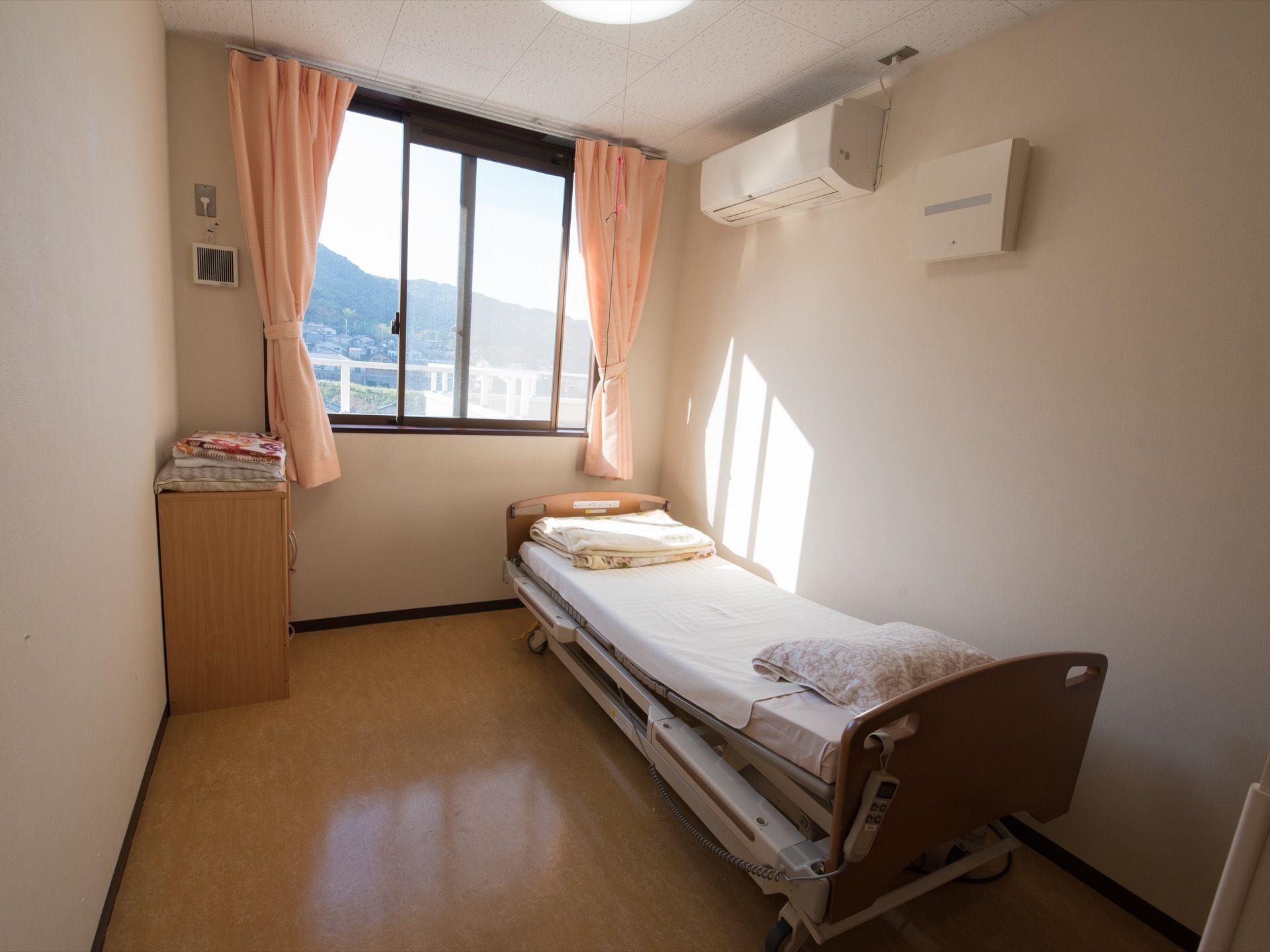 白色の壁に茶色の床の居室である。正面に窓があり、壁際にベッドを置いている。天井下には、エアコンの設備を整えている。