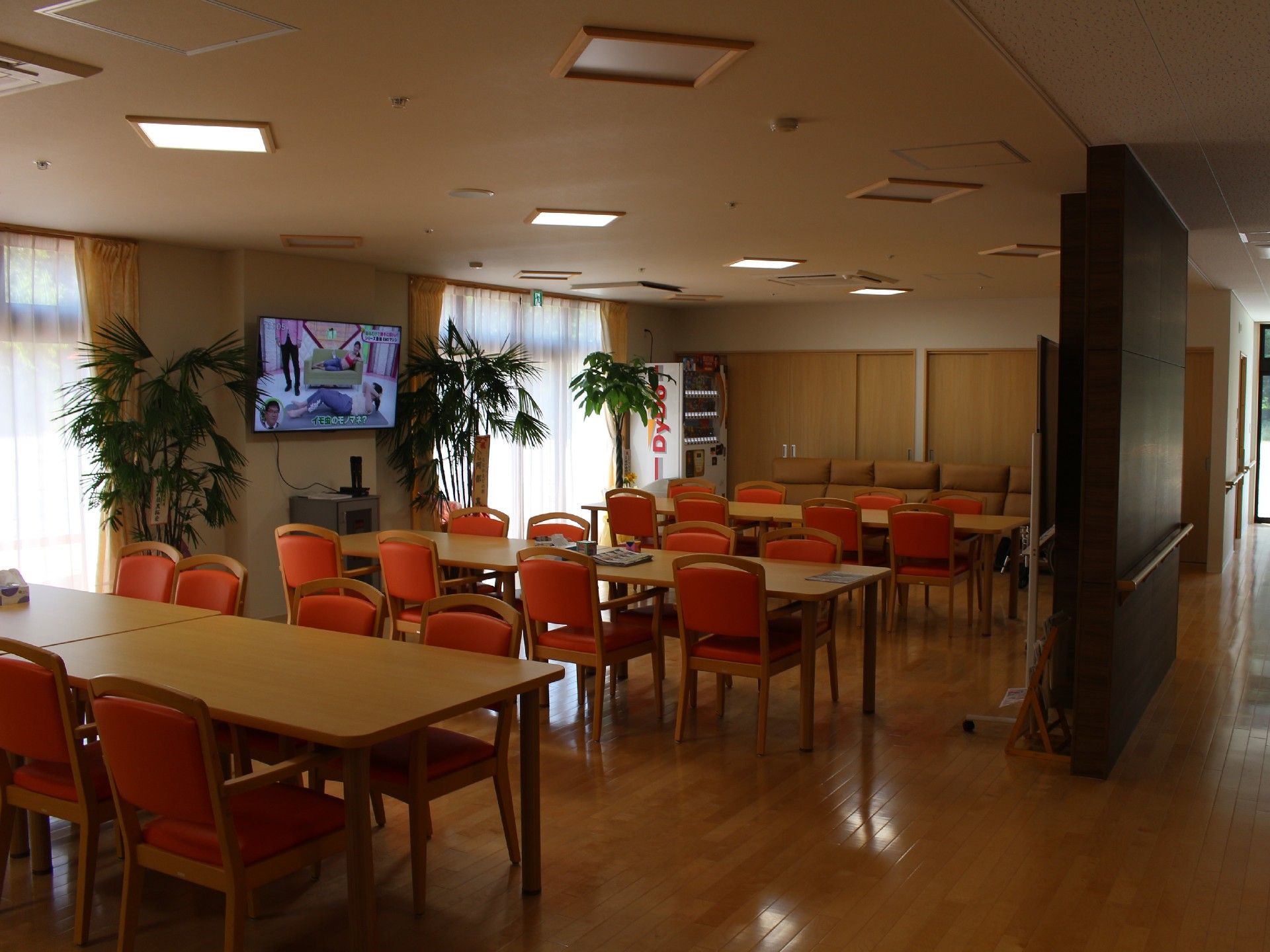 テレビや自動販売機が設置された広いホール内には、入居者様が楽しい時間をお過ごしいただけるよう、テーブルと椅子が置かれている。