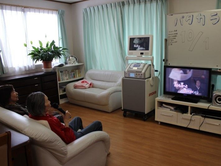 カラオケ設備が整ったリビングで入居者様が歌を楽しんでいる。室内にはテレビやソファ、ホワイトボードなどが置かれている。