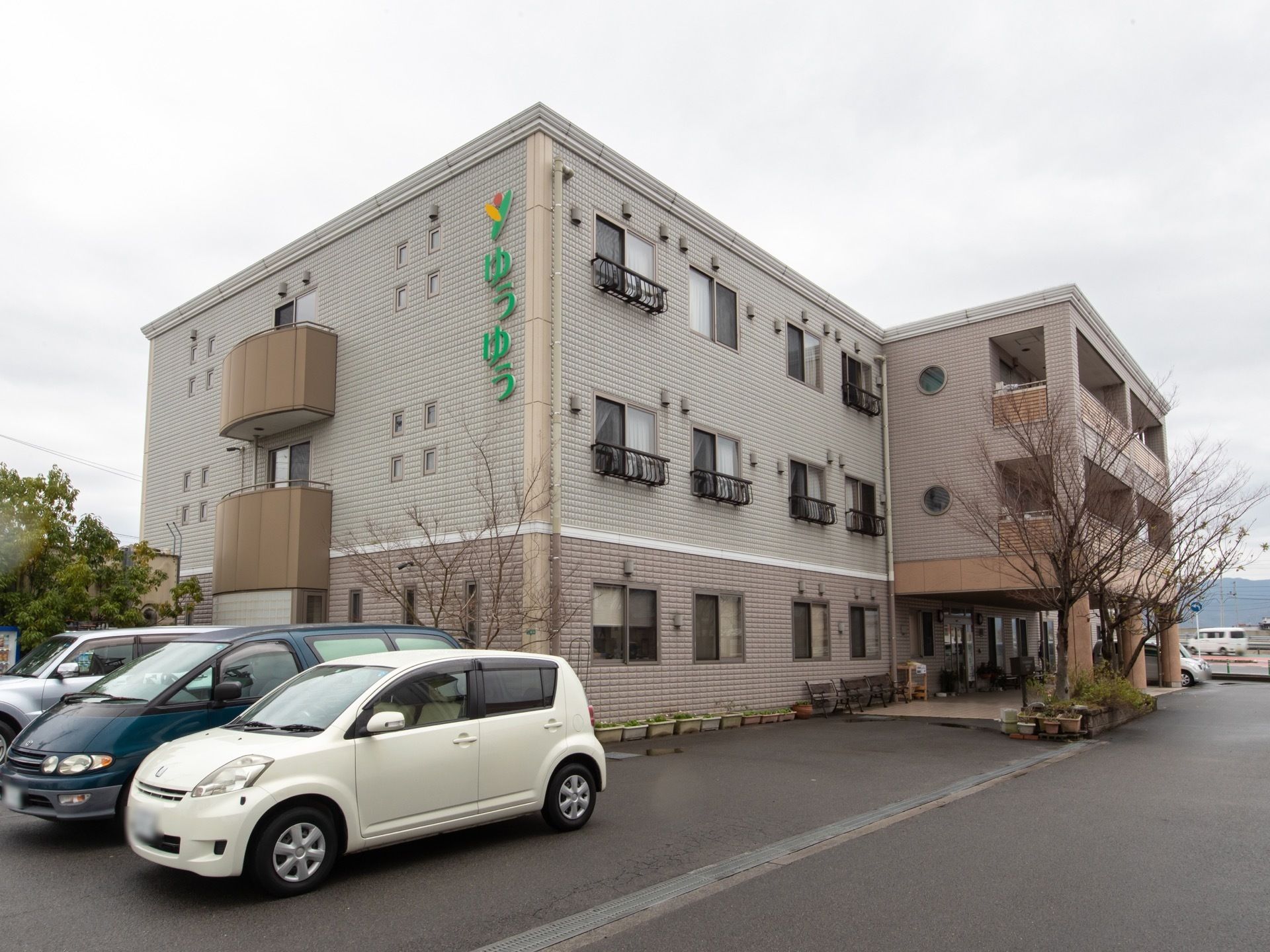 ブラウン色の上品なデザインの外観である。建物の横には駐車スペースがあり、玄関の前にはいくつかベンチが置かれている。