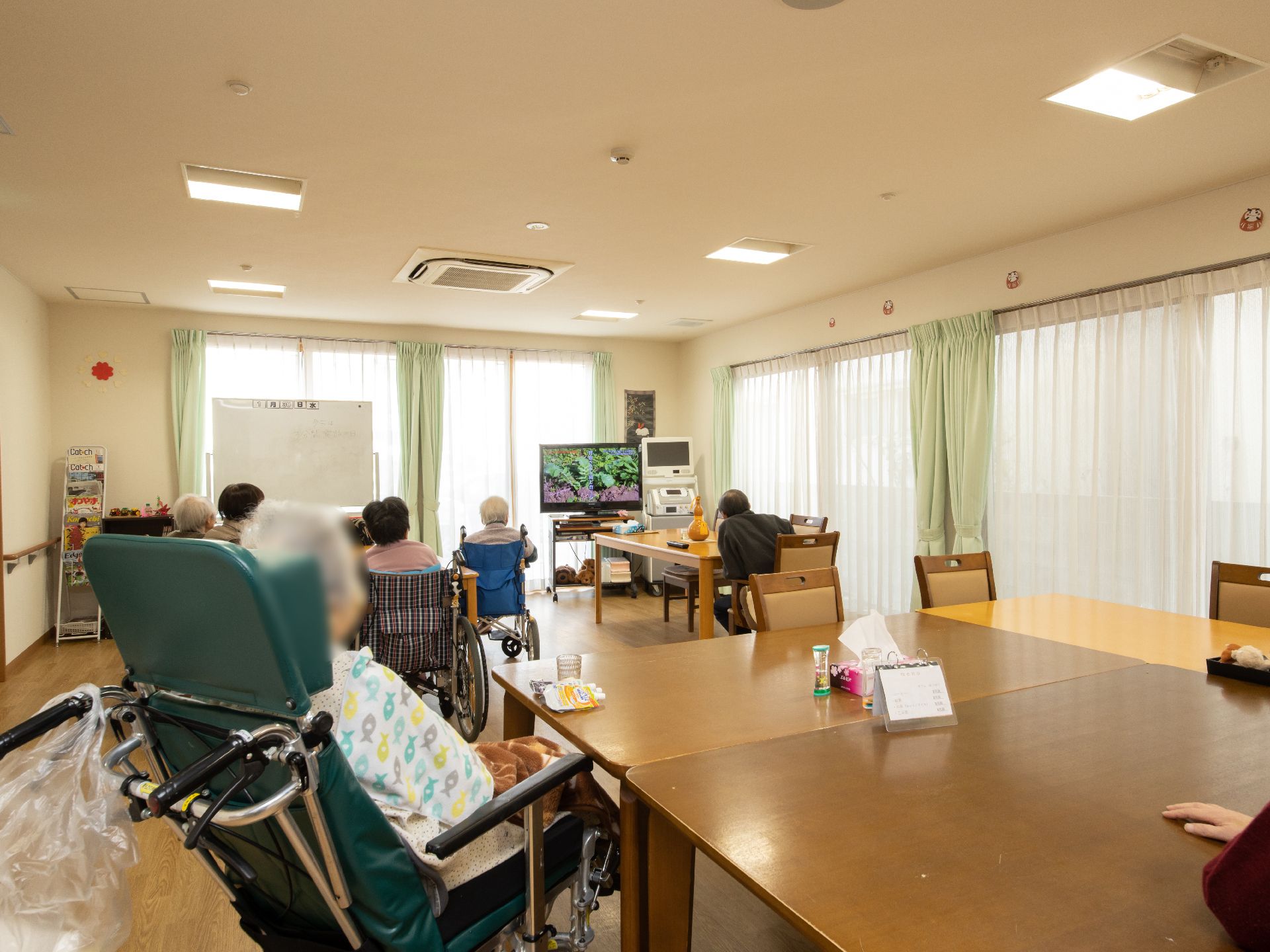 広い食堂内に数名の入居者様が集い、奥にある大型テレビを鑑賞している。車椅子をご利用の入居者様が多く見える。