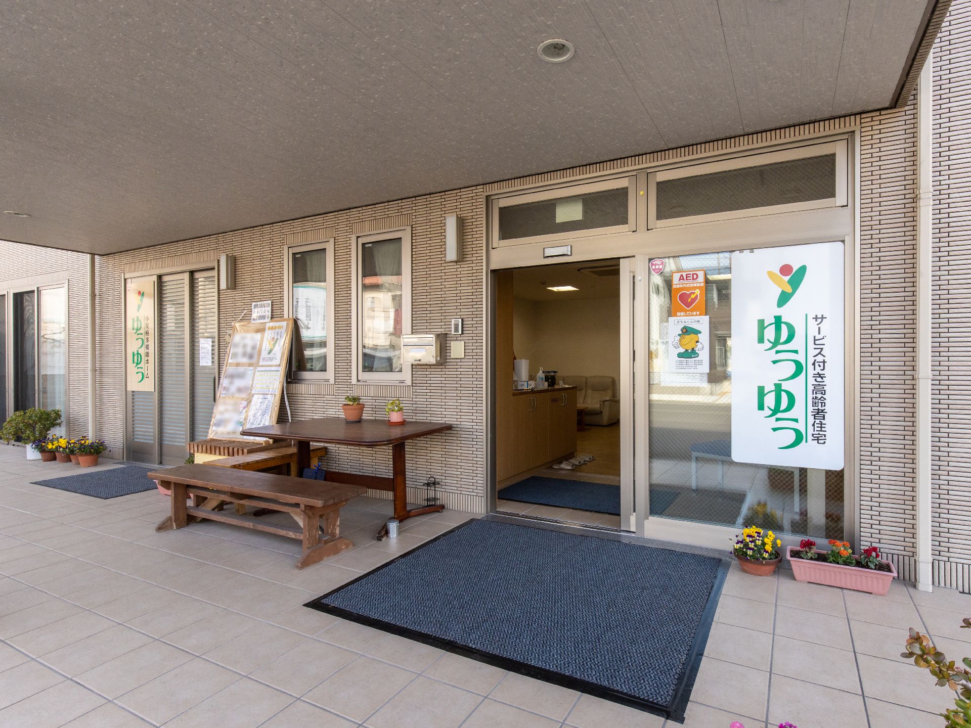 玄関は自動ドアになっており、横には木製のテーブルとベンチが置かれている。ドア前にはプランターが並んでいる。