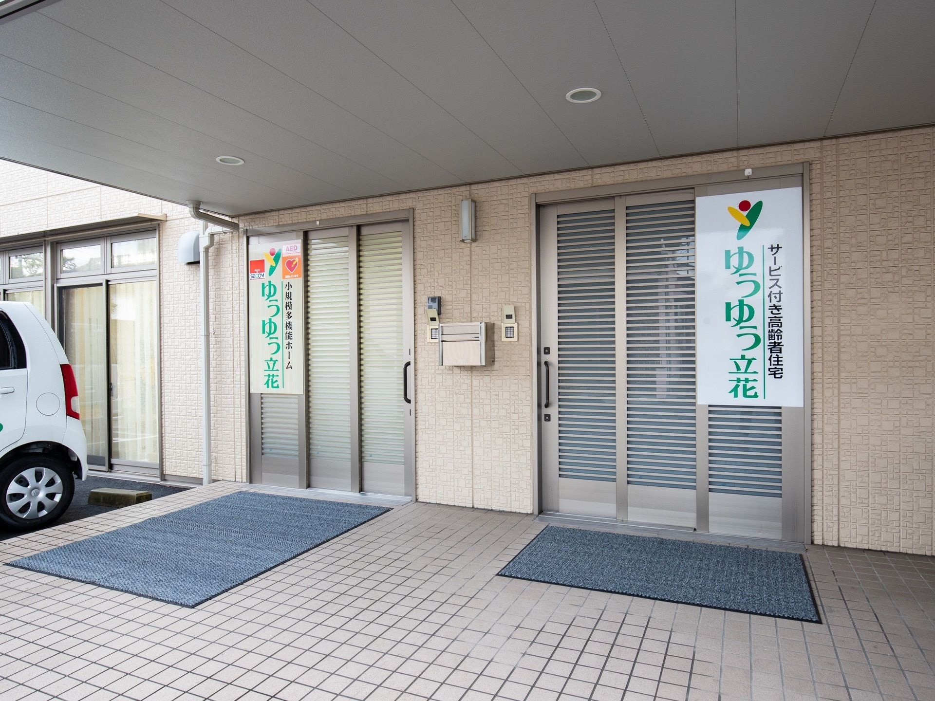 ベージュ色のタイル張りのポーチに、二箇所扉がついている。それぞれの扉に看板があり、手前にグレーのマットが敷かれている。