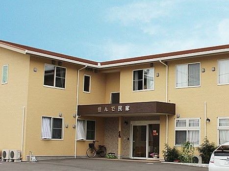 民家のような三角屋根の建物で、各所に窓があり、いくつかは開いている。建物の前はアスファルト舗装されており、白い車が1台停まっている。