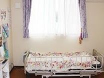紫色のカーテンの付いた窓の下に介護用ベッドが置かれている。左の壁際にポットののったサイドテーブルが見える。