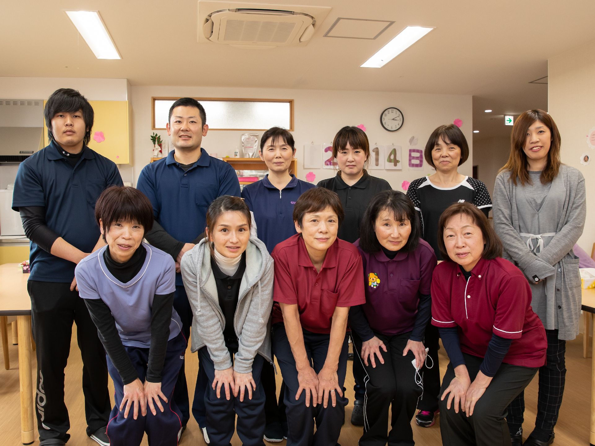 男性2名、女性9名のスタッフがポロシャツやスポーツウェアを着て、共有スペースで整列している。にこやかに微笑んでいる。