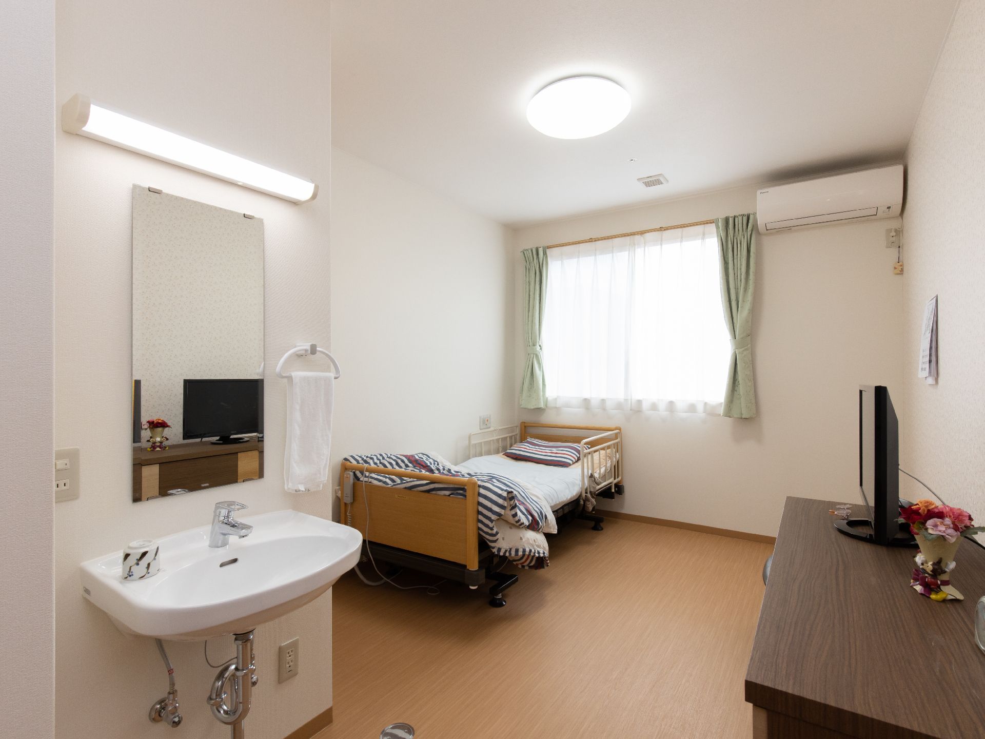 居室の壁は白、床はフローリング敷になっている。窓際に電動式ベッドを置いている。入口付近には洗面台が設置されている。