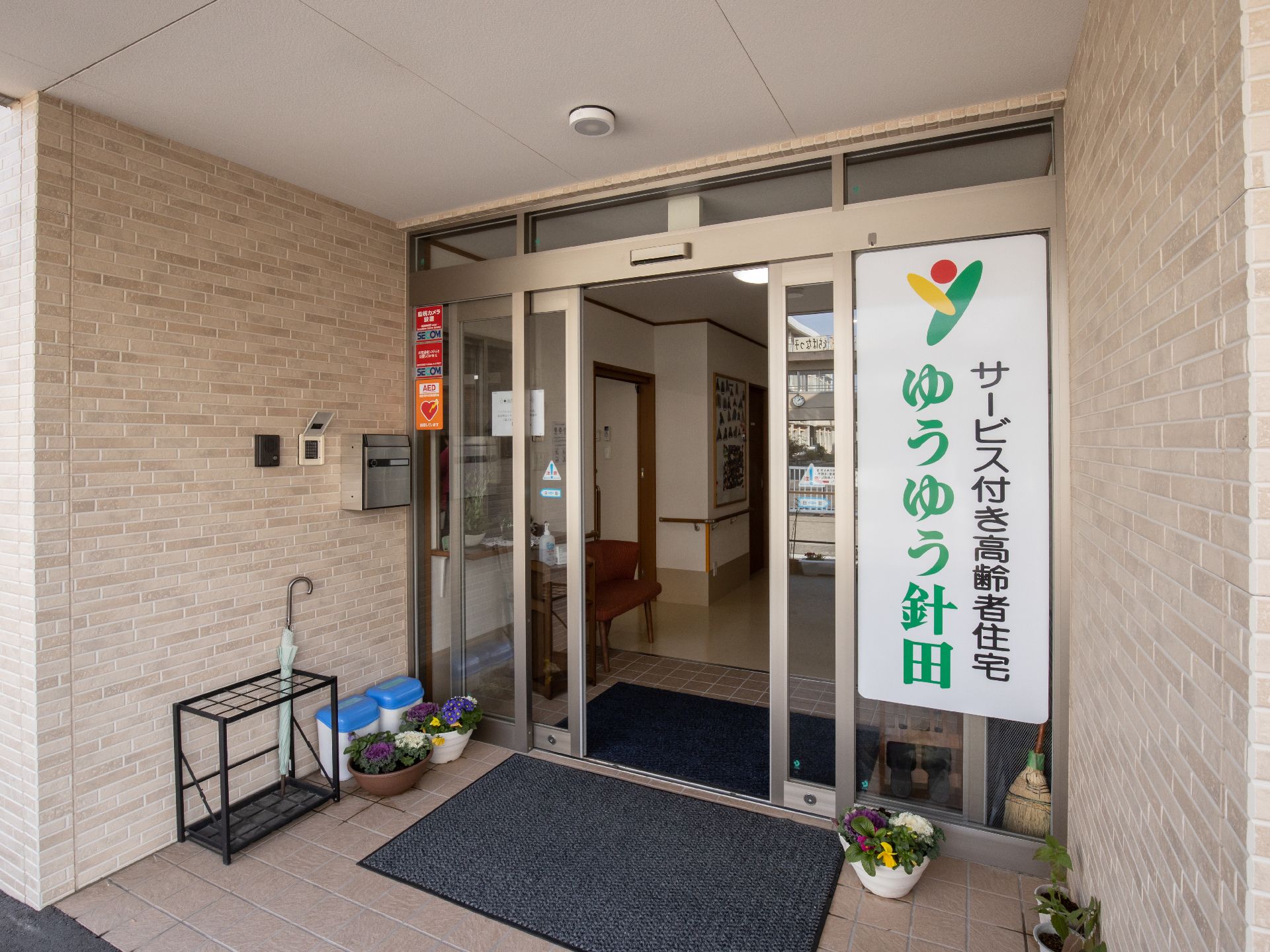 両開きのガラスの自動扉の入口である。入口の右側に大きな看板があり、施設名とロゴが描かれている。入口前に傘立てがある。