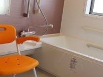 浴室の浴槽側には窓がある。浴槽内には横型の手すりが設置されている。洗い場にはオレンジ色の介護用シャワーチェアーが用意されている。