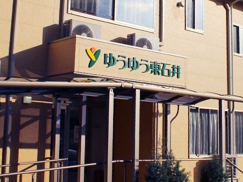 薄い茶色い外観の建物がある。屋根の下の通路両側には、手すりが取り付けられている。屋根の上には施設名が書かれている。室外機や窓もある。