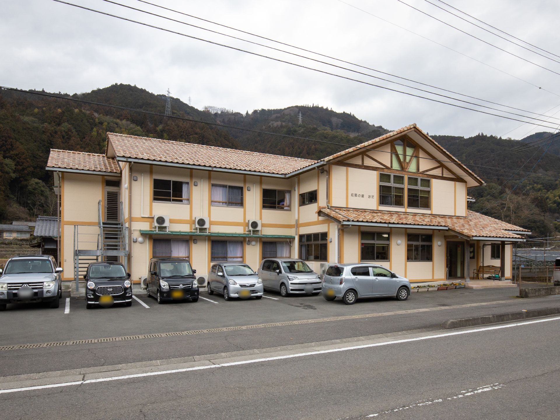 屋根はレンガ風の色合いで、壁は明るいクリーム色の２階建てのおしゃれな施設である。建物の前が駐車場になっている。