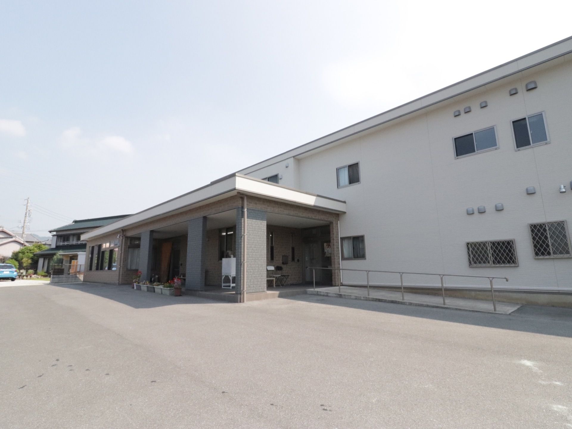 広い敷地に２階建ての施設建物が建っている。玄関口周辺は大きなひさしになっていて、前にはプランターが飾られている。