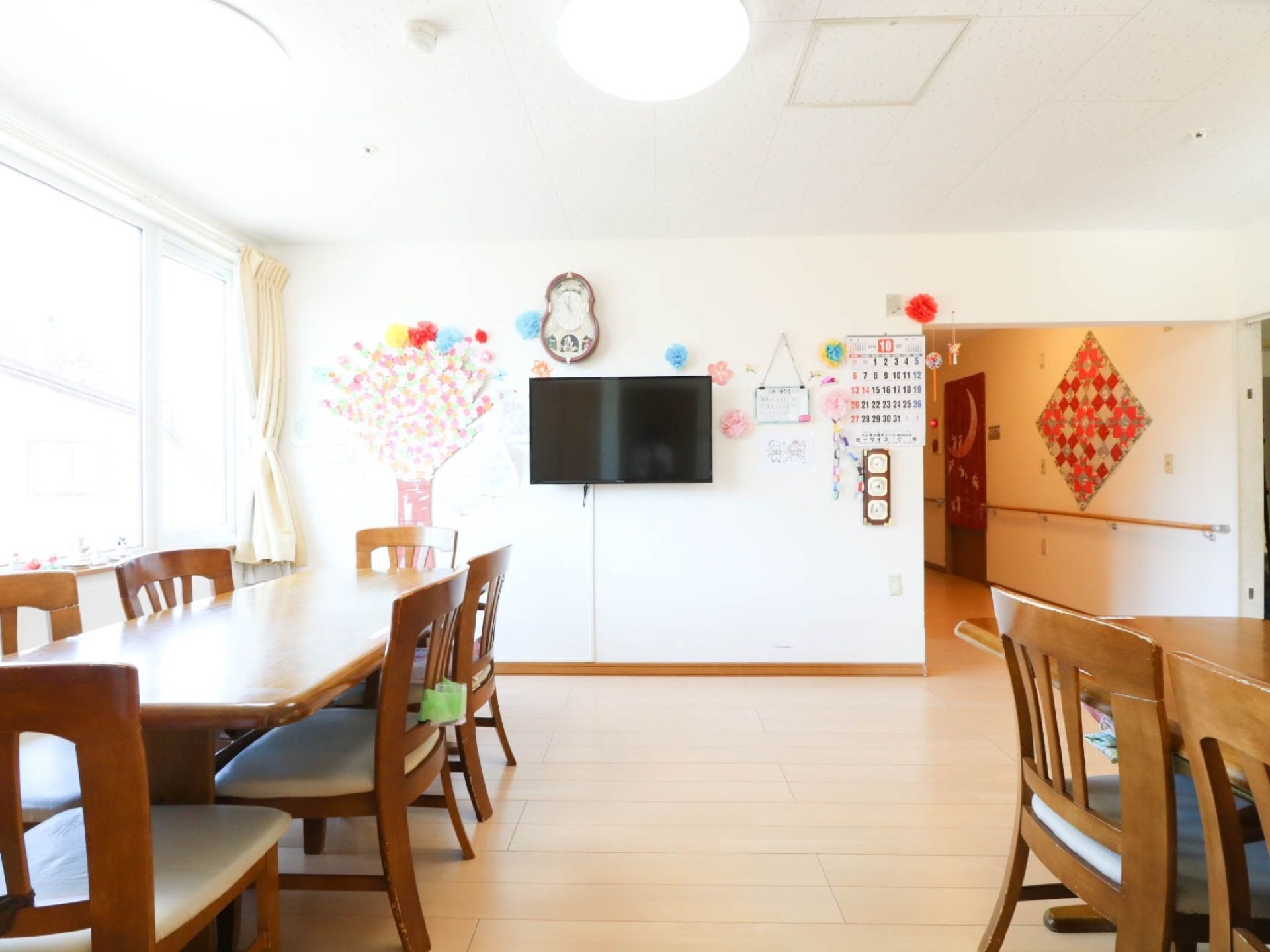 壁に、時計や大型テレビが掛けられたフロアがある。ダイニングテーブルが並べられており、窓から陽が入って明るくなっている。