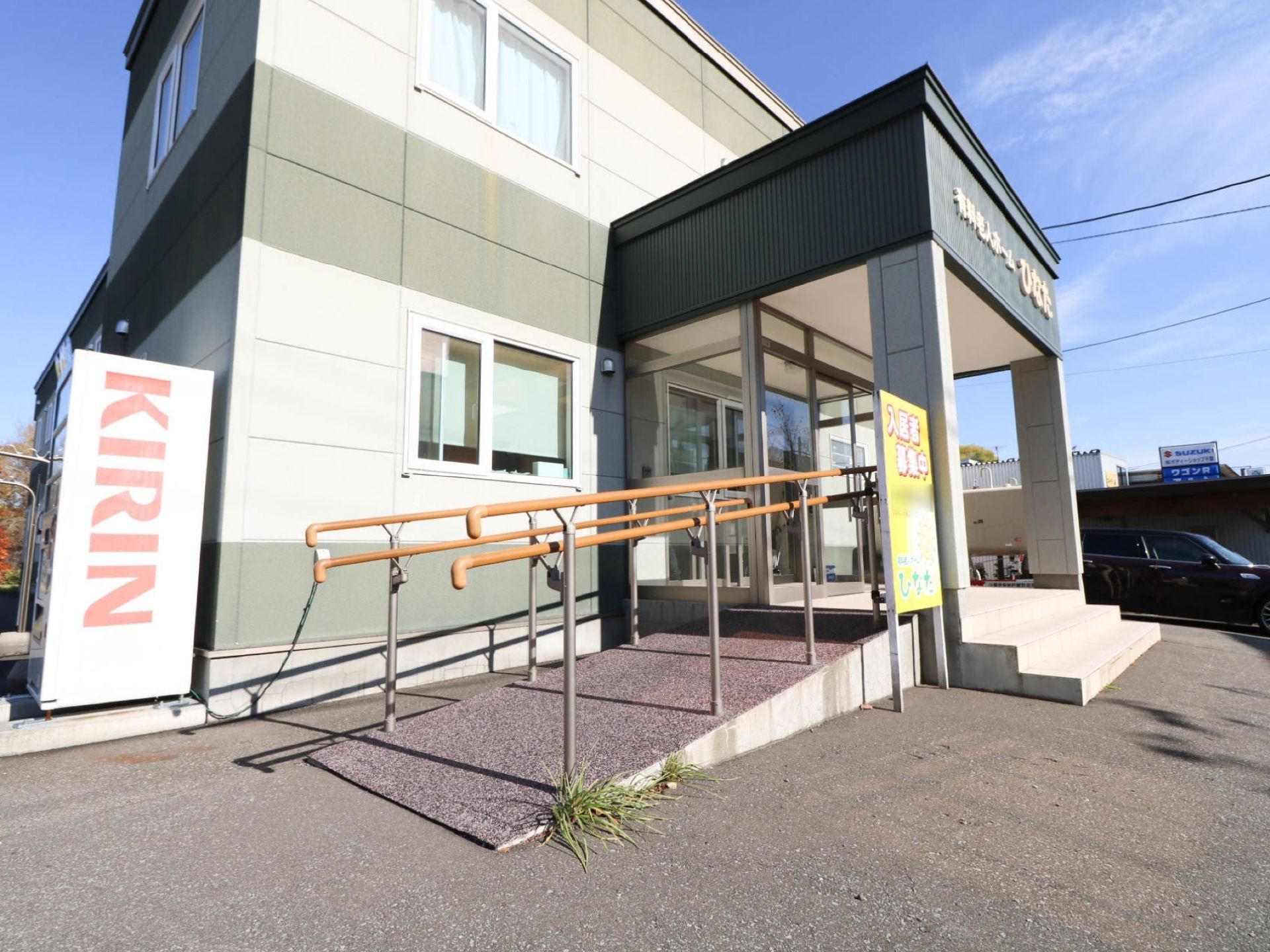 ２階建ての住宅があり、壁際に自動販売機が置かれている。玄関の横には、木目調の手すりが付いたスロープが設置されている。