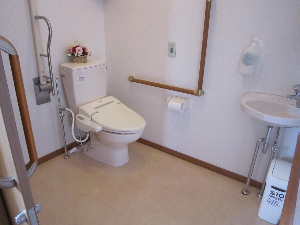 ゆったりとしたスペースのトイレである。便座の横側の壁にエル型の手すりを備え、前方にシンプルな洗面台を設置している。