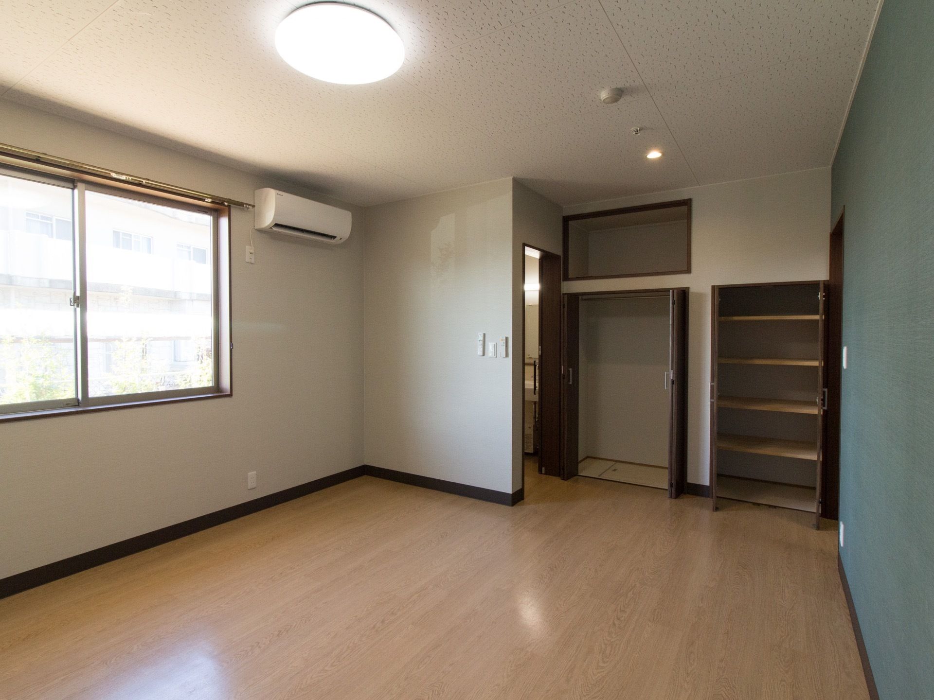 窓付でフローリングの一室。白い壁なので、窓から陽の光が入り明るく見える。トイレの横に収納棚が複数設置してある。