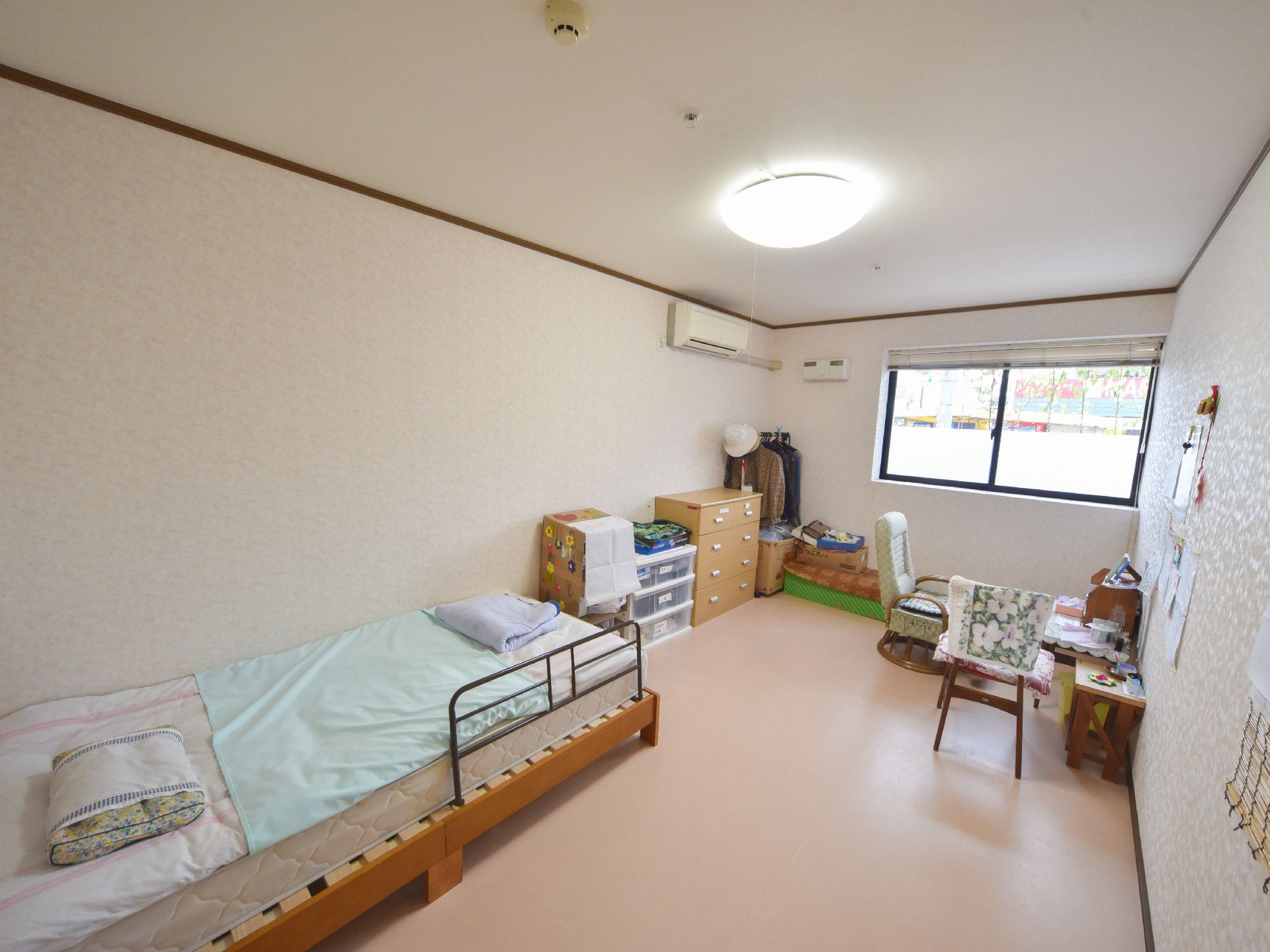 ピンク系の床が明るい雰囲気の広々とした居室にはベッドや椅子、チェスト等が置かれている。大きな窓から光が差し込んでいる。
