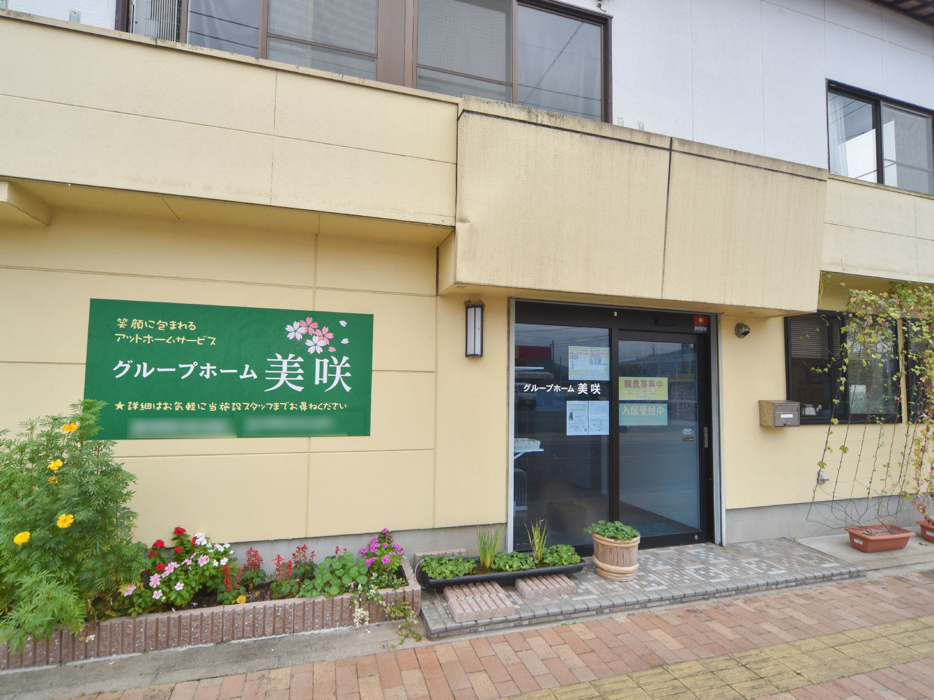 黄色の外壁で、緑地の横長の看板が取り付けられている玄関付近