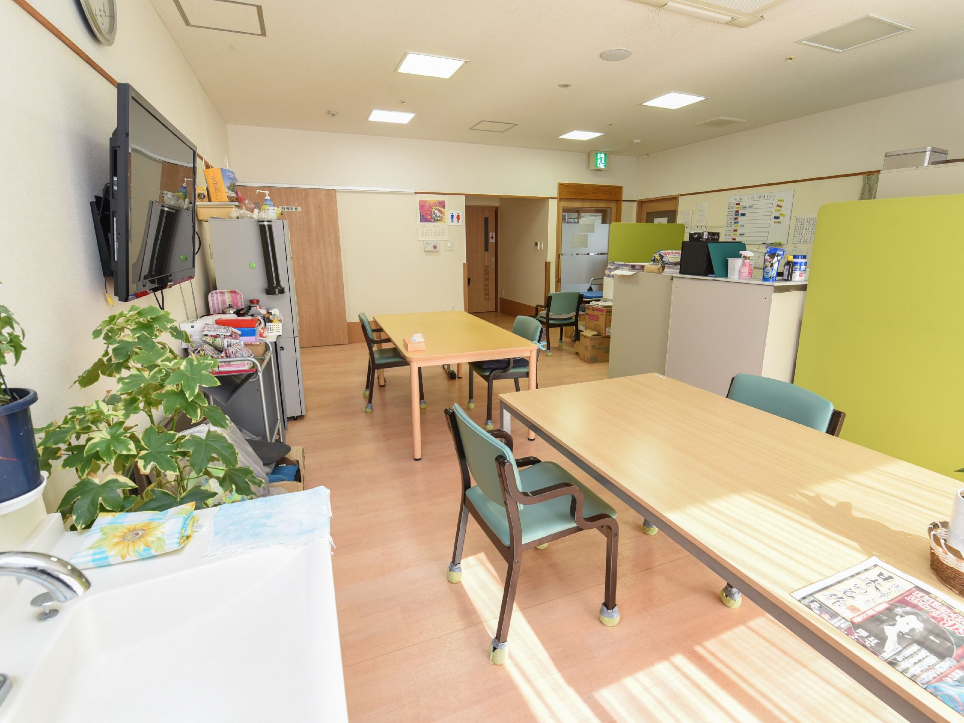 中央に横長のテーブルが設置されており、新聞紙やティッシュ箱などが置かれている。壁に黒いテレビが掛けられている。