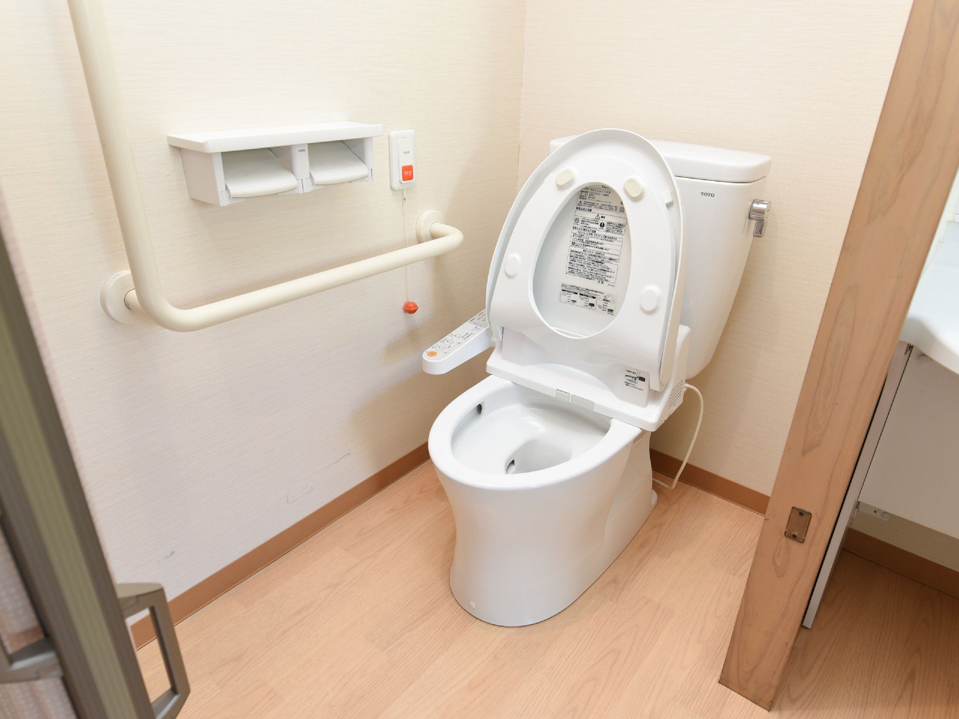 白い壁とフローリングの床の個室トイレで、壁にL字型の白い手すりが取りつけられている。ペーパーホルダーの右側にナースコールがある。